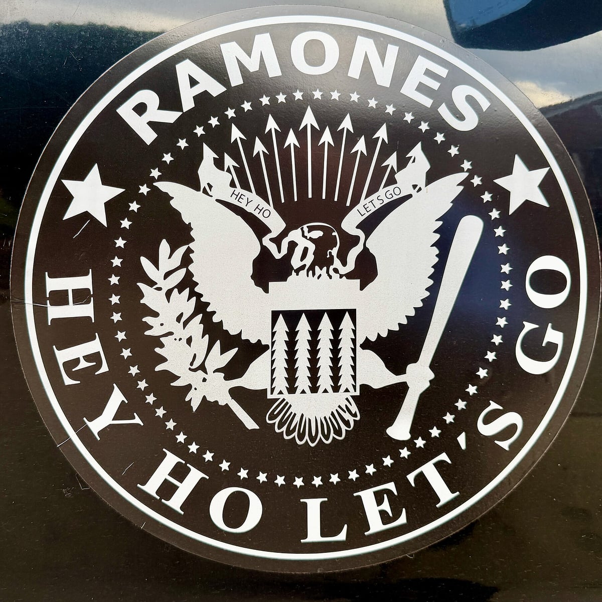Black and white Ramones seal decal with an eagle holding arrows and a baseball bat, and the words Hey Ho Let's Go around the rim