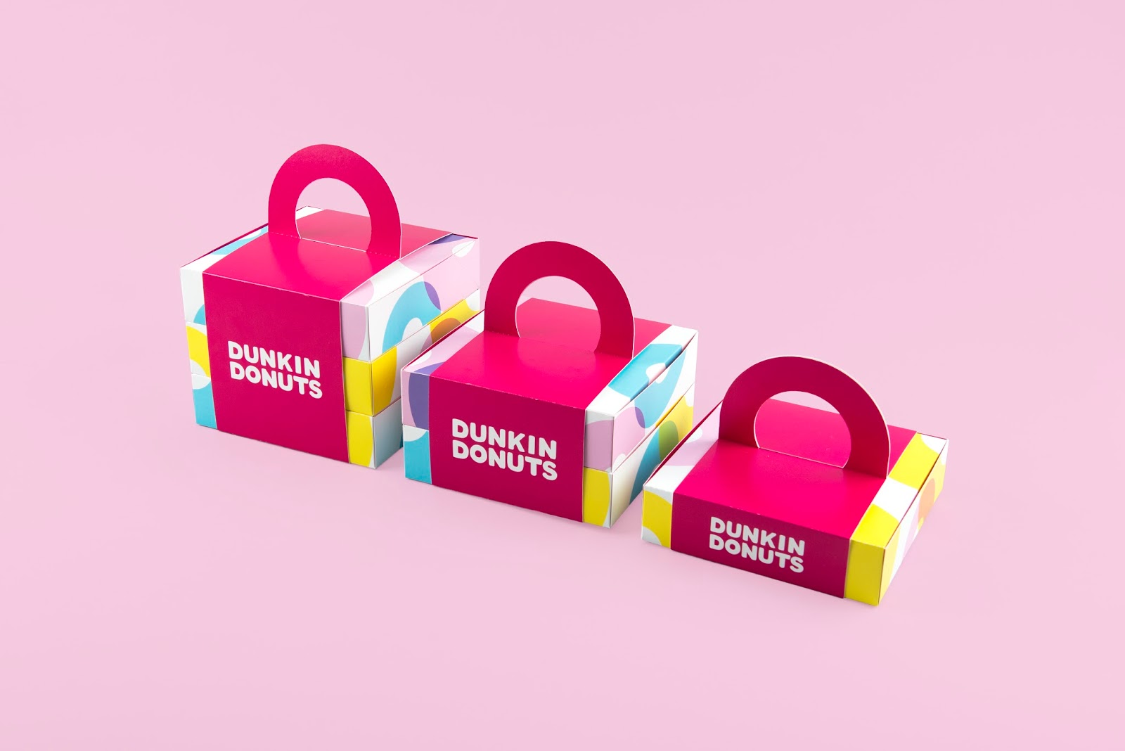 Three hot pink donut boxes with Dunkin Donuts printed on the side and yellow, white and blue panels, on a light pink background